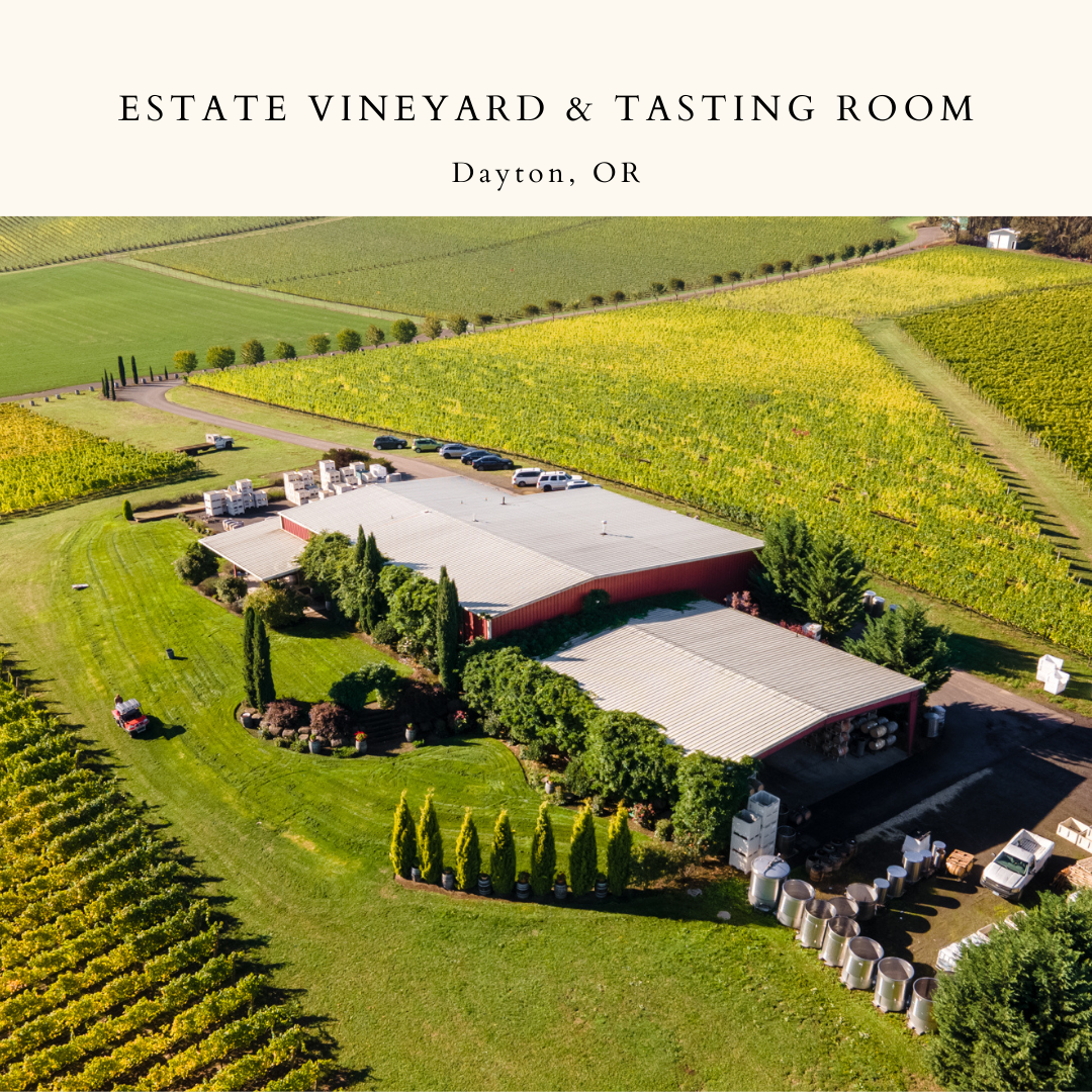 An aerial view of the Estate Vineyard and Tasting Room. The building is surrounded by evergreen trees and a luscious lawn. Behind the building, some cars are seen parked in front of the estate vines.