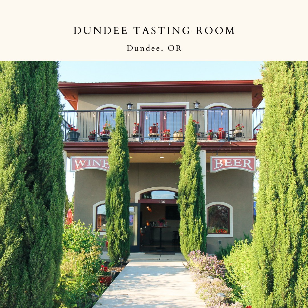 A frontal view of our Dundee Tasting Room. A sidewalk leads to the front door, surrounded on both sides by evergreen trees and flowers. On either side of the building, signs advertise 