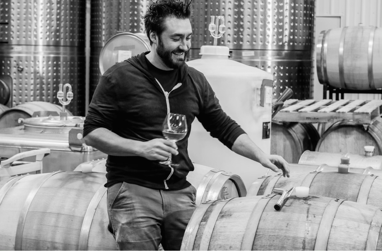 Our winemaker, Greg McClellan, holds a glass of wine next to several barrels of wine. He is reaching for the barrel in front of him, looking away from the camera.