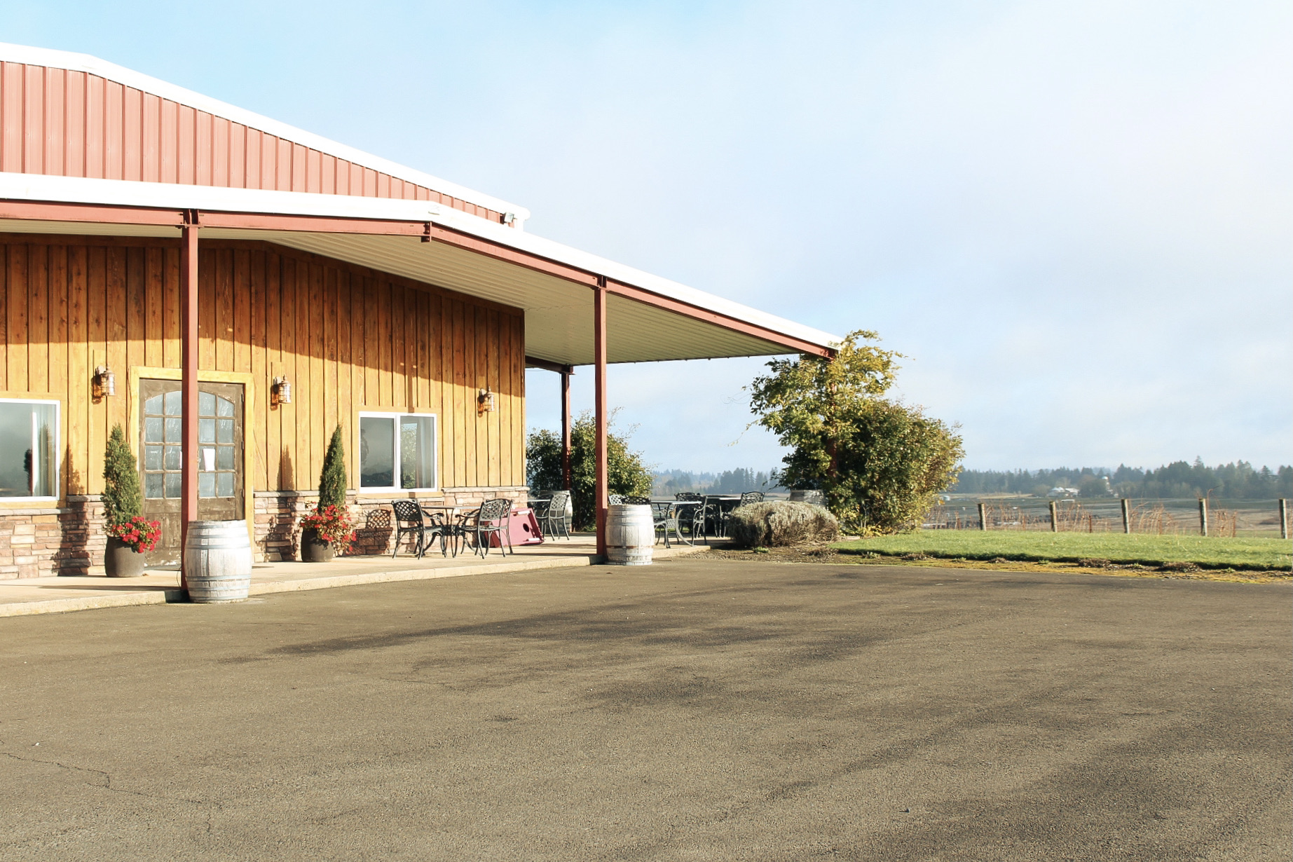 A frontal view of our Estate Tasting Room, with small potted evergreens on either side of the double doors, and several sets of outdoor tables and chairs fill the covered patio to the right of the building. In the distance to the right, the beginnings of several rows of vines can be seen.